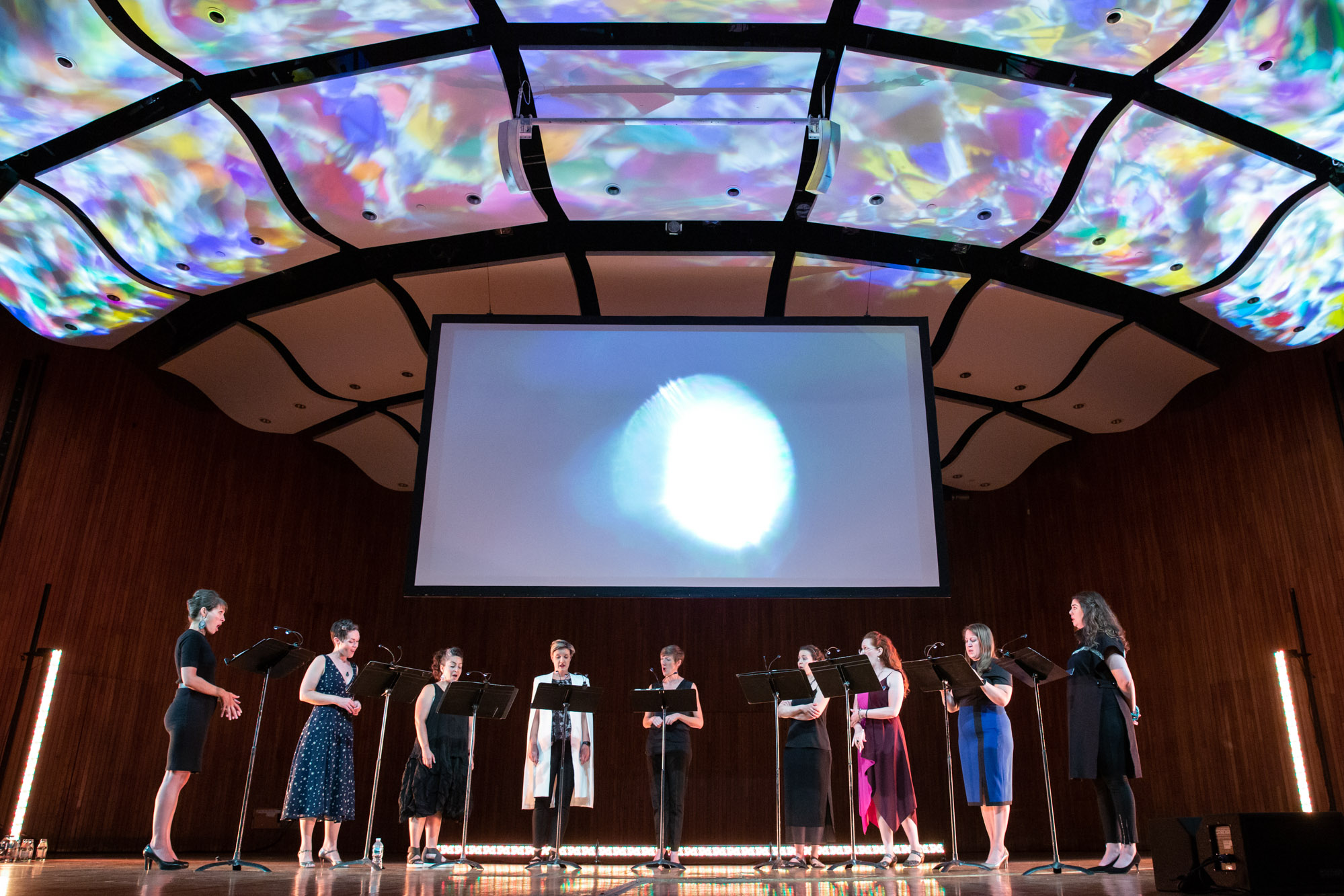 Nine women (lorelei ensemble) sing on a stage while multicolored projections float on the ceiling and a moon-like shape is projected on a screen behind them
