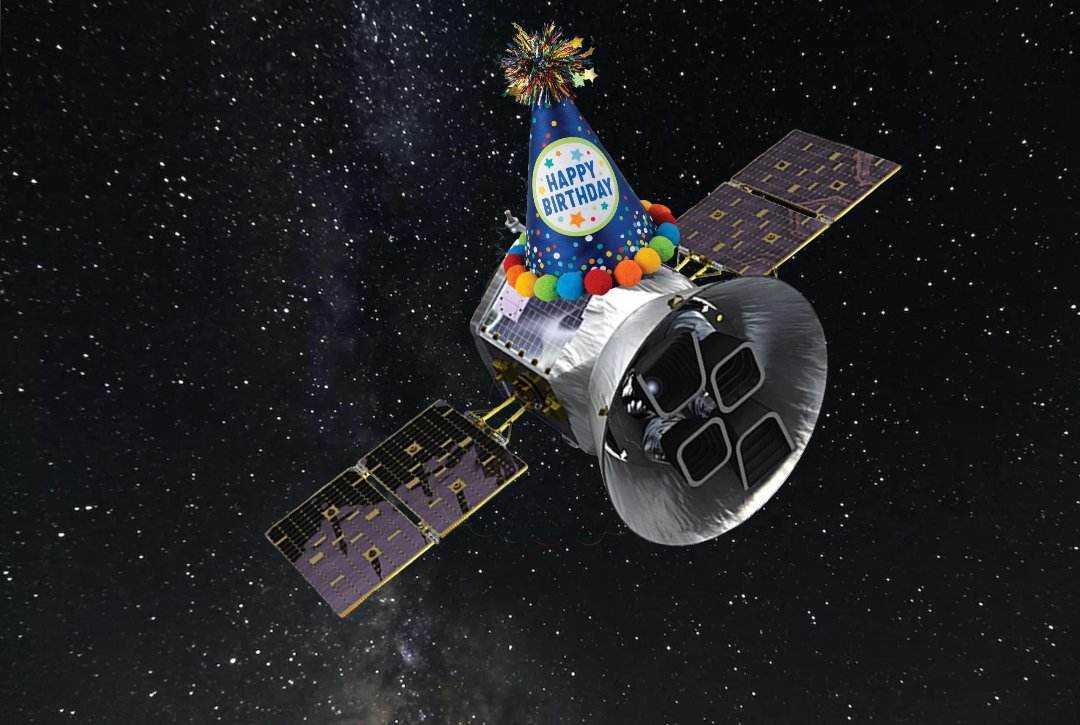 An illustration of the TESS spacecraft against a starry background with a pointed birthday hat perched on top of the spacecraft.